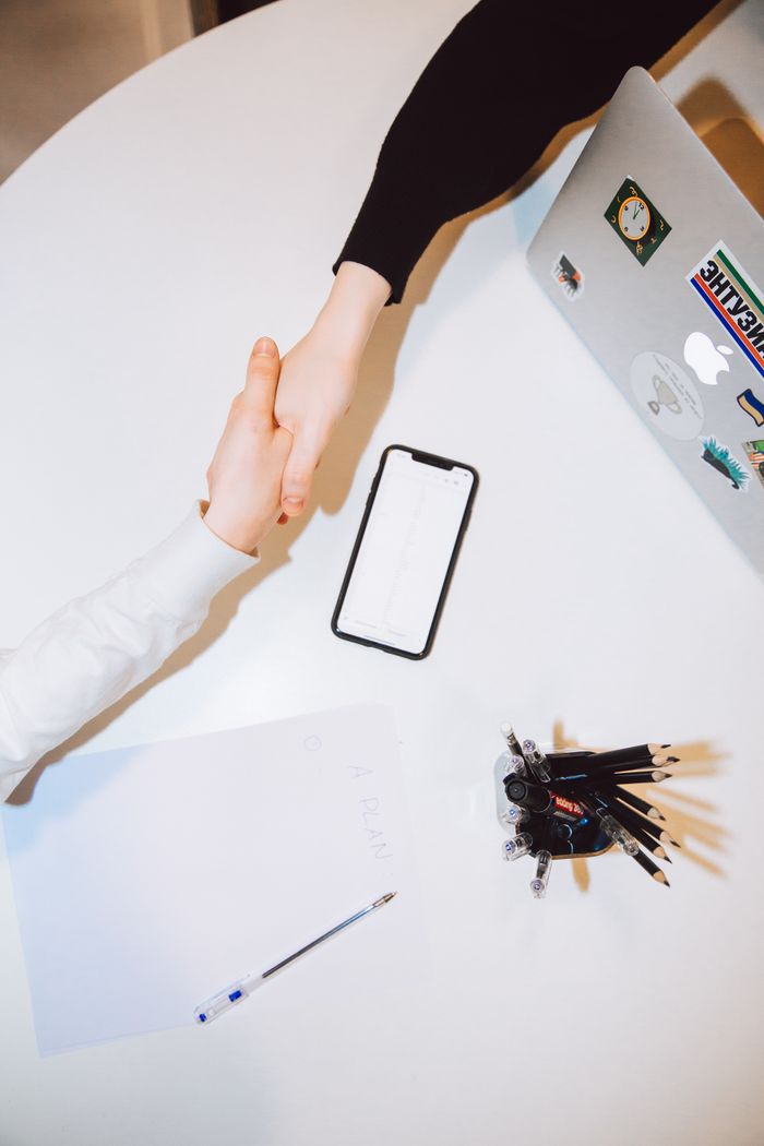 vision Two individuals shaking hands across a desk with a smartphone and laptop in a modern office setting.